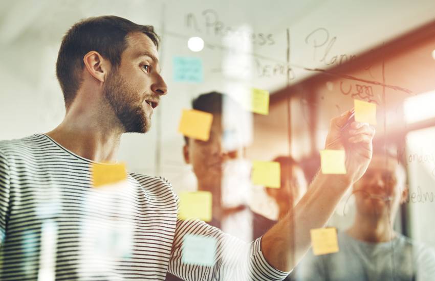 Group of casually-dressed businessmen in a brainstorming session utilizing stick notes and writing on a glass dry-erase board.