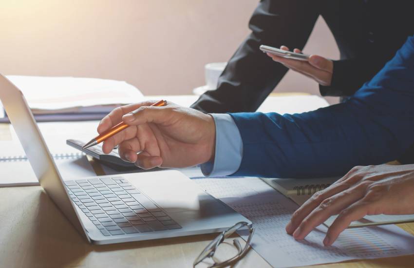 Close up of two businessmen reconciling the contents of a printed report with their laptop.