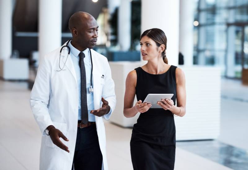 Female medical sales rep holds a tablet and speaks with a doctor in the lobby of a hospital