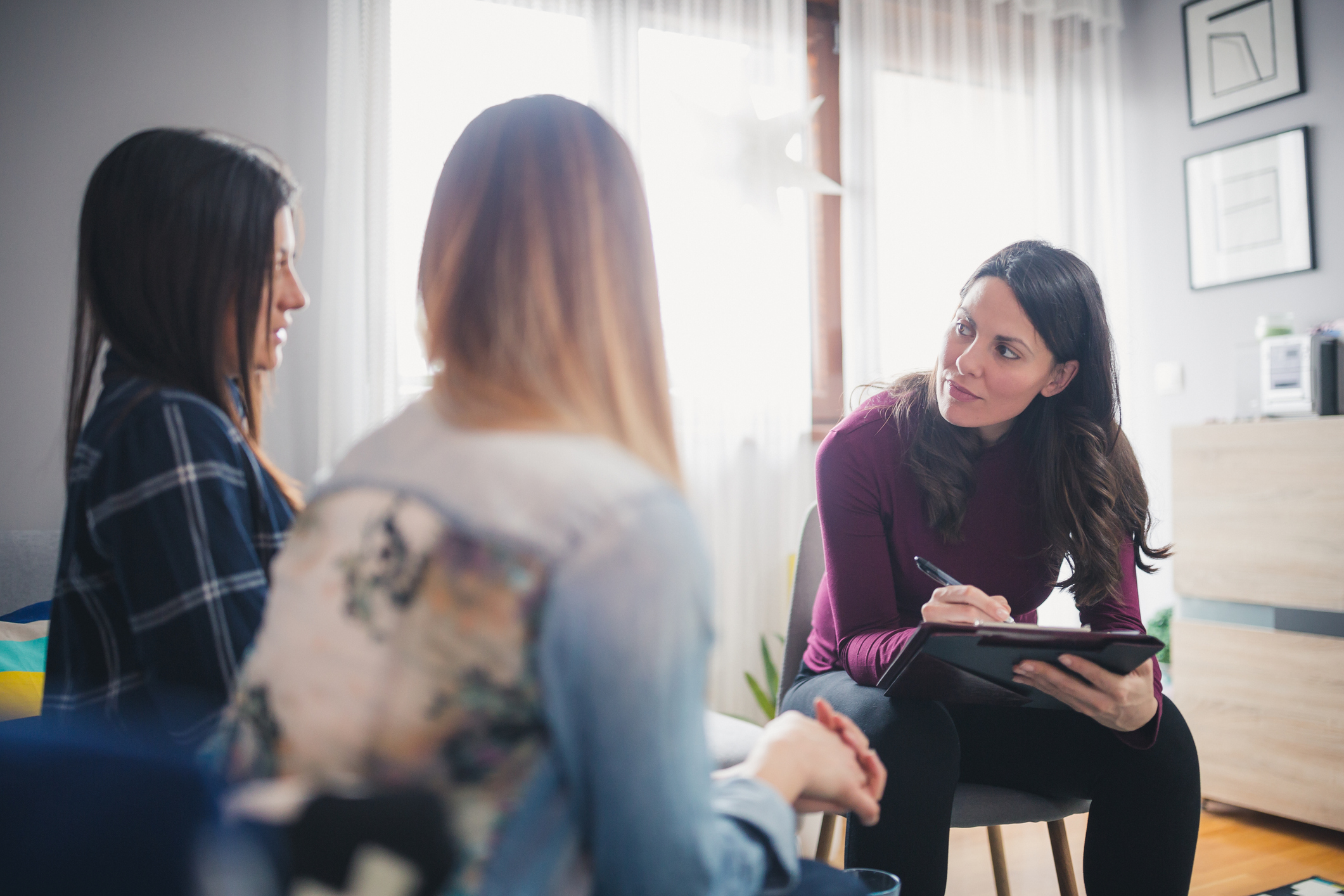 Female therapist listens intently to two female patients and takes notes on her clipboard.