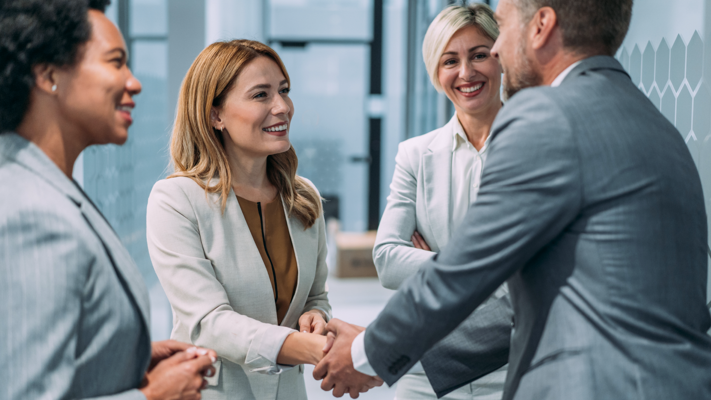 Four business professionals in suits, two women and two men, are shaking hands and smiling in a modern office setting.