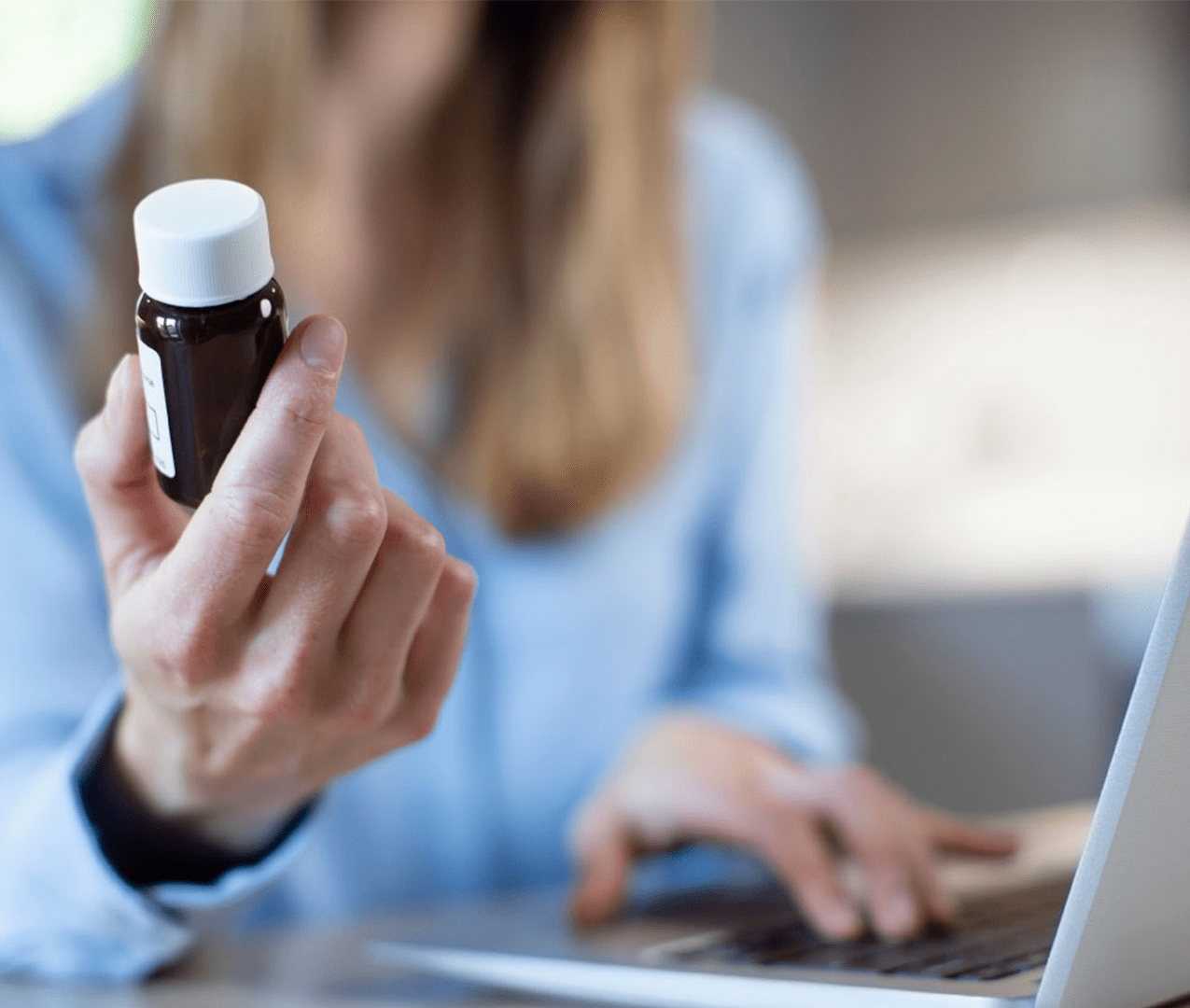 Woman holding a bottle of medicine, looking at the label as she types on a laptop.