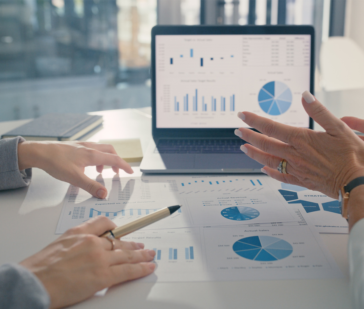 Close up of female hands seemingly in discussion over several report documents full of charts and graphs and a laptop with a reporting dashboard in the background
