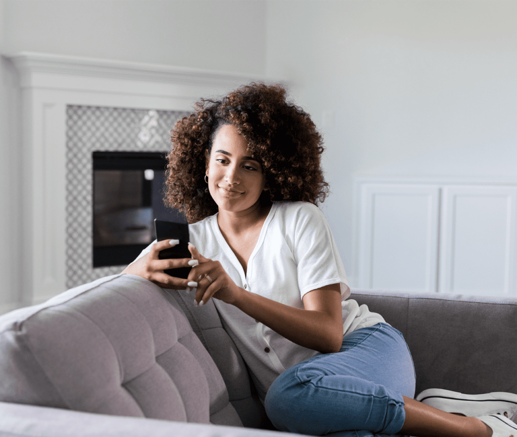 Young woman sits on her couch and texts with her cellphone