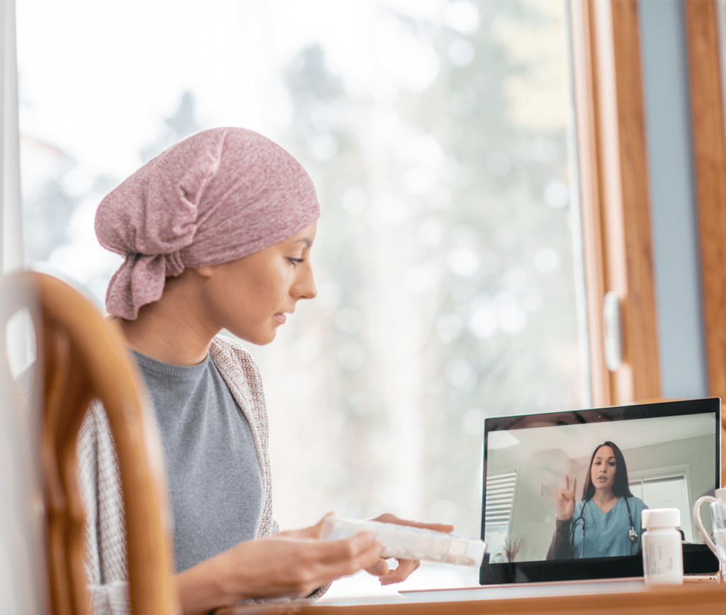 Young woman with her head wrapped in a scarf holding a weekly medicine organizer and talking to her doctor via telemedicine on her laptop.
