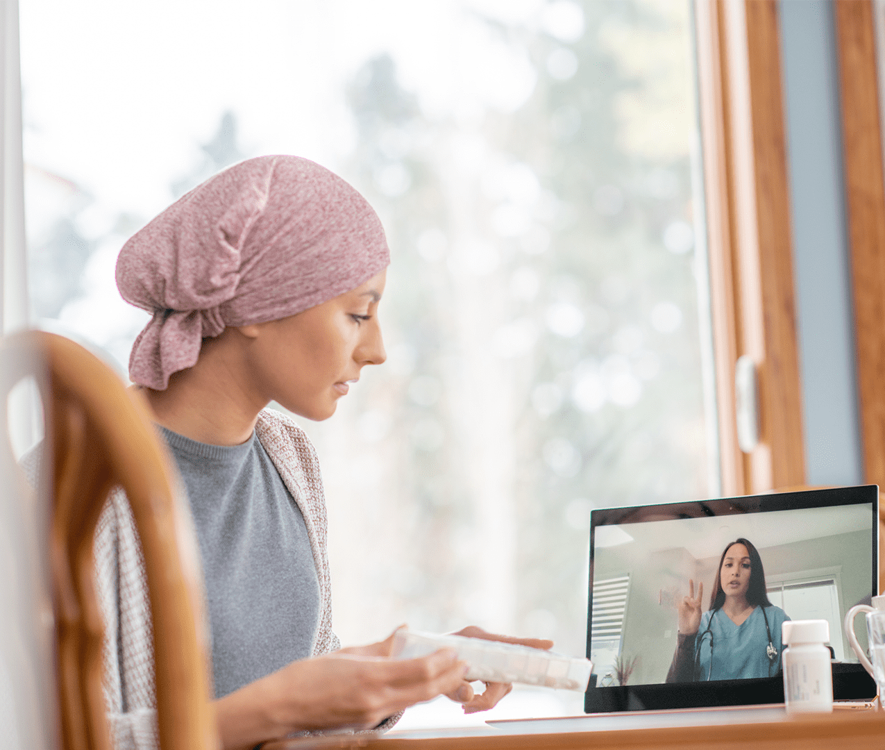 Young woman with her head wrapped in a scarf holding a weekly medicine organizer and talking to her doctor via telemedicine on her laptop.