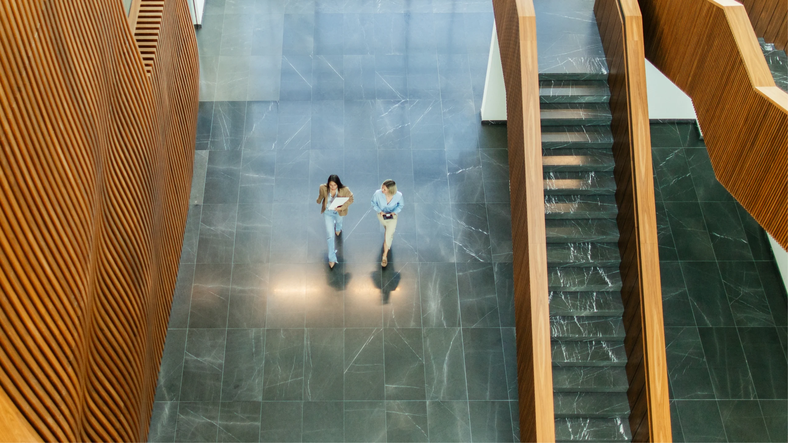 Two women walk across a large, modern lobby with dark marble floors, flanked by wooden slatted walls and stairs.