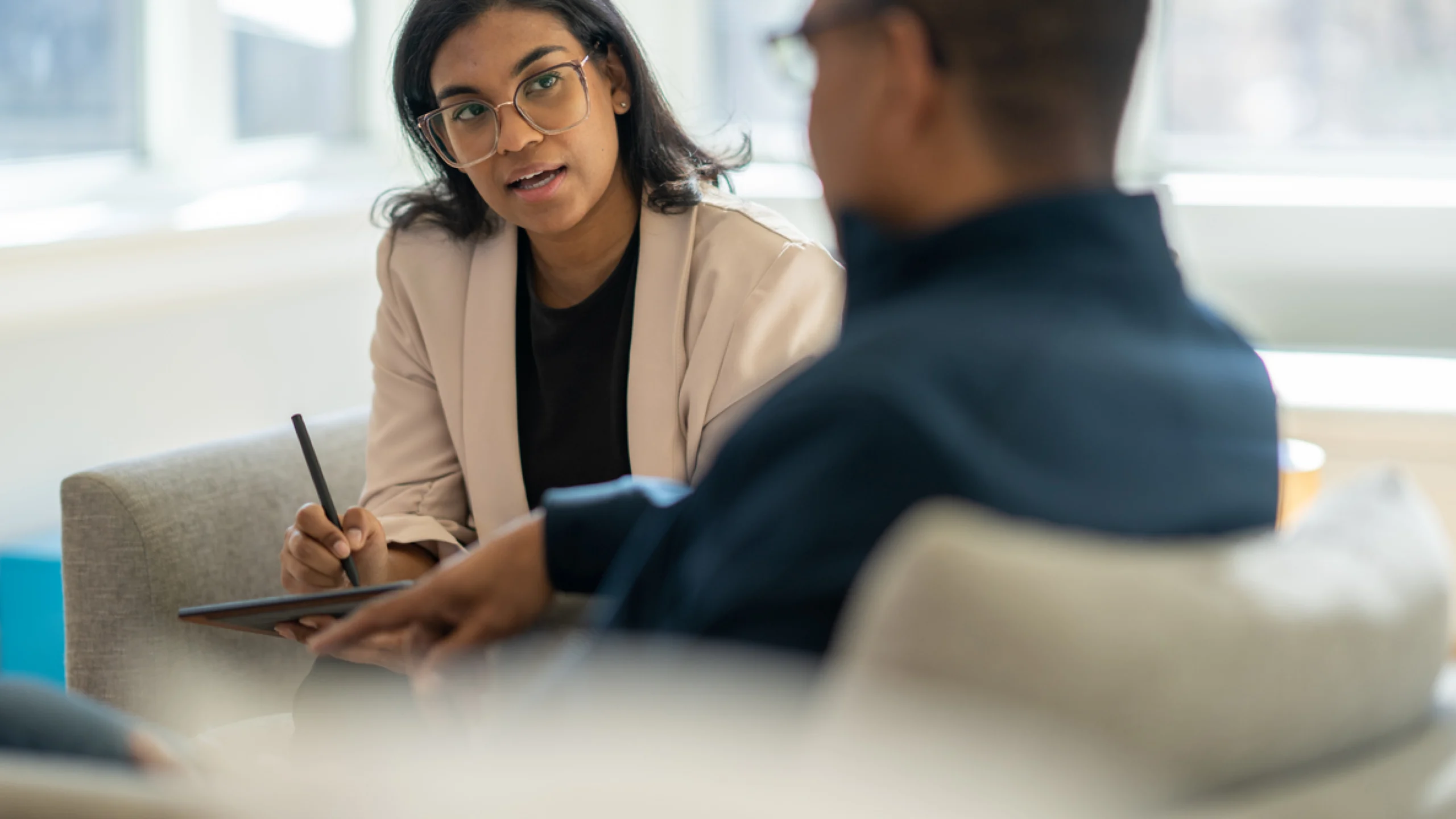 A woman in glasses and a blazer speaks to a person, writing on a tablet during a consultation or interview.