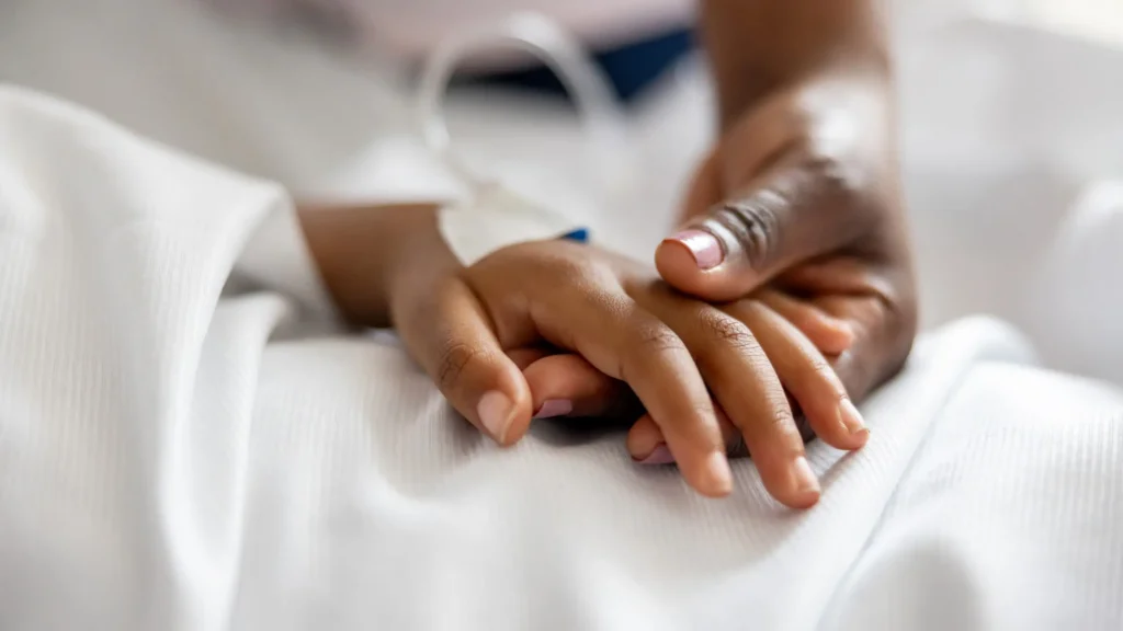 A close-up of two hands, one gently holding the other, resting on white bedsheets, conveying comfort and support.