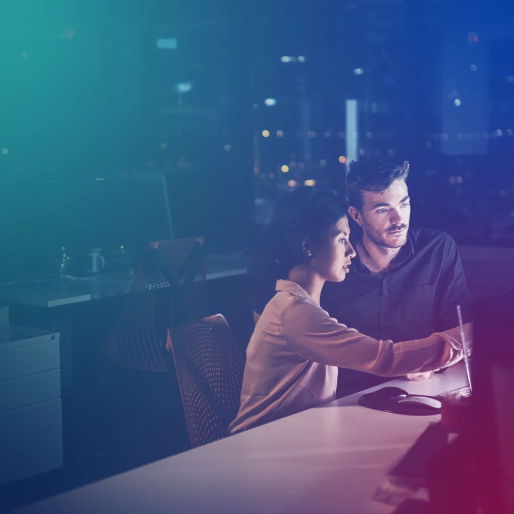 Two colleagues, a man and a woman, working together at a desk in a dimly lit office at night, looking at a computer screen.