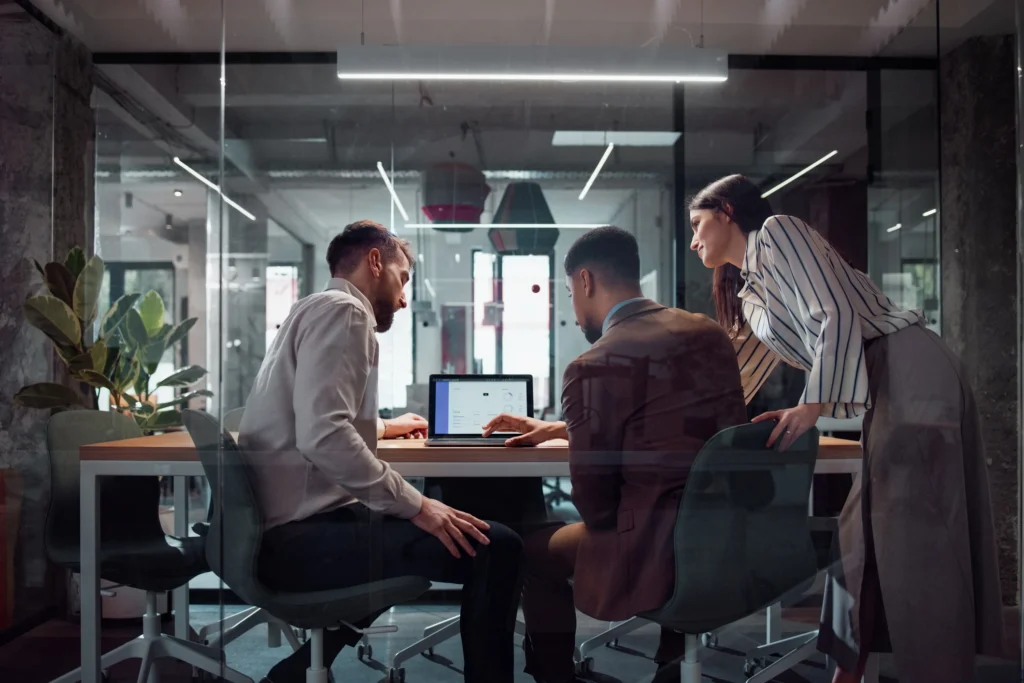 Three business professionals, two men and one woman, collaborate around a laptop displaying charts and data in a modern office.