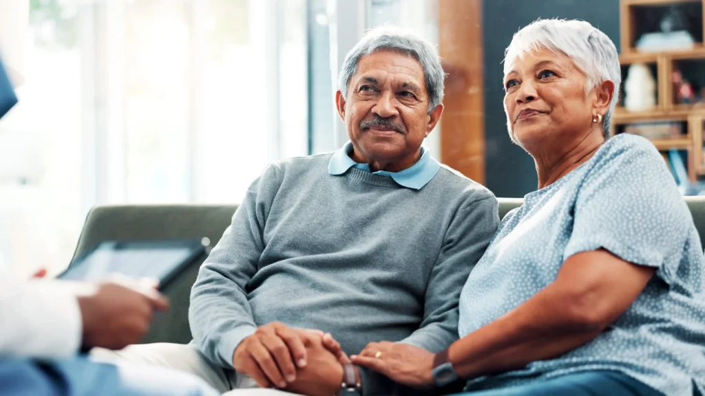 An elderly couple, a man and a woman, are seated on a couch, looking attentively towards someone off-camera. The man has a mustache and gray hair, wearing a gray sweater. The woman has short white hair and is wearing a light blue patterned top. Their hands are clasped together, suggesting comfort and support.