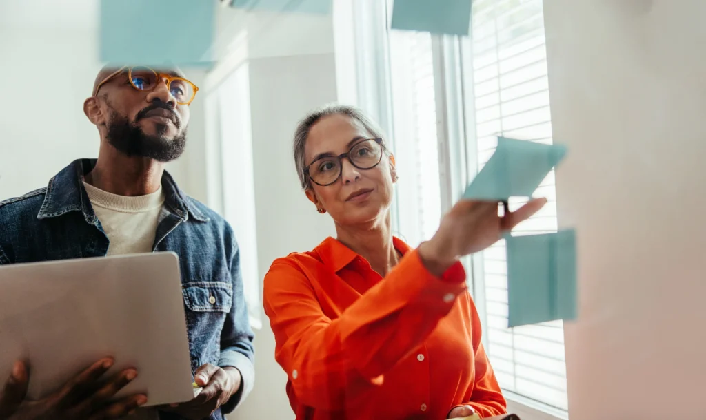 Two diverse colleagues collaborating in an office, with one placing a sticky note on a glass board and the other holding a laptop.